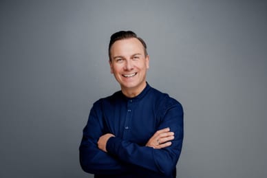 das-excellence-gourmetfestival-4.jpg Lächelnde Person in blauer Kleidung mit verschränkten Armen vor grauem Hintergrund.