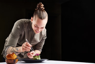 das-excellence-gourmetfestival-10.jpg Person in Kochjacke verziert ein Gericht auf einem Tisch.
