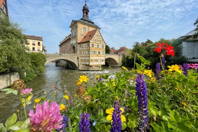 fruehlingsfrisch-donau-und-franken-2.jpg Fachwerkgebäude auf einer Brücke über einen Fluss, umgeben von bunten Blumen.