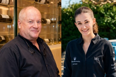 das-excellence-gourmetfestival-10.png Zwei Personen in dunkler Kleidung lächeln in die Kamera. Im Hintergrund sind Weinflaschen und draussen Sitzgelegenheiten zu sehen.