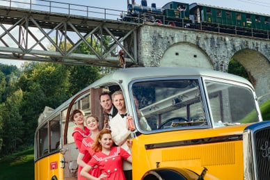 sing-und-swingauf-dem-rhein-1.jpg Gruppe in 1950er-Jahre Kleidung vor gelbem Oldtimer-Bus mit Eisenbahnbrücke im Hintergrund.