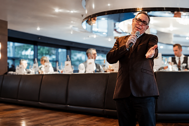 an-die-kueste-der-normandie-1.png Person im Anzug singt in einem Restaurant, umgeben von sitzenden Gästen.