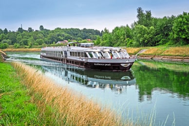 Das Flusskreuzfahrtschiff Excellence Pearl gleitet auf einem ruhigen Fluss durch eine grüne Uferland­schaft.