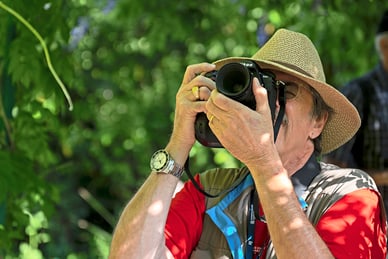 mit-der-kamera-von-der-seine-erzaehlen-3.jpg Mann mit Hut fotografiert lila Blumen im Garten.