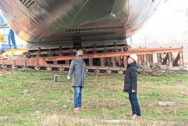 Zwei Arbeiter mit Schutzhelmen betrachten ein grosses zylindrisches Bauteil auf rotem Transportgestell auf dem Werftgelände.