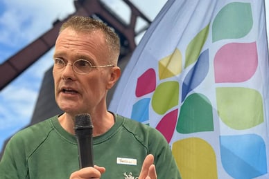 lebensraeume-der-zukunft-iga-2027-1.jpg Person hält Mikrofon und spricht neben einer farbenfrohen Flagge und Stahlkonstruktion.
