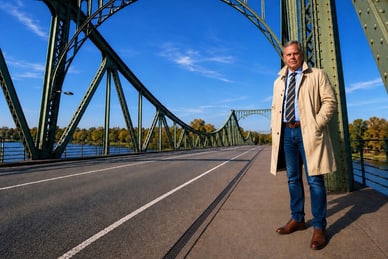 nordsommer-havelland-heide-hanse-3.jpg Person in beigem Mantel steht auf einer Brücke mit markanten Metallbögen.