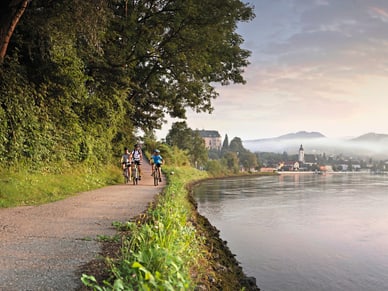 Drei Radfahrer auf gepflastertem Uferweg an der Donau bei Grein mit Kirchturm im Morgendunst.