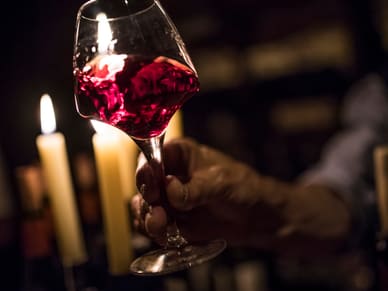 Eine Hand hält ein Glas mit tiefrotem Wein, im Hintergrund leuchten zwei Kerzen sanft.