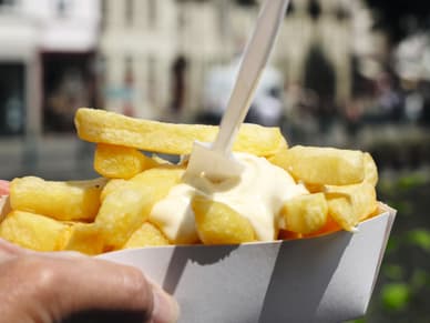Eine Hand hält einen Papierbehälter mit belgischen Frieten und cremigem Dip vor einer sonnigen Altstadtkulisse in Gent.