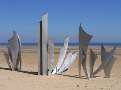 Abstrakte Edelstahlskulpturen des Denkmals Les Braves am Sandstrand von Omaha Beach unter blauem Himmel.