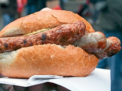 Eine Hand hält ein Brötchen mit gegrillter Bratwurst und brauner Sosse, im Hintergrund verschwommene Gebäude.