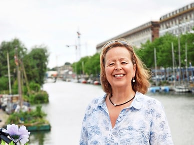 Elsbeth Gugger lächelt neben einem lila Fahrrad mit Blumen am Lenker, im Hintergrund ein Amsterdamer Kanal.
