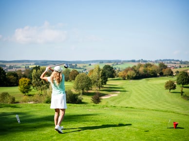 Frau in hellgrünem Shirt und weissem Rock führt einen Golfschlag auf einem weitläufigen Golfplatz aus.