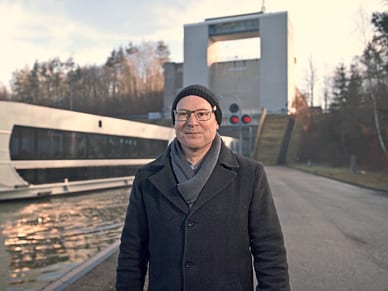 Mann in Winterkleidung steht auf gepflastertem Weg neben einem modernen Schiff am Flussufer, grosses Bogengebäude im Hintergrund.