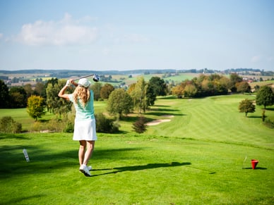 Frau in hellgrünem Shirt und weissem Rock führt einen Golfschlag auf einem weitläufigen Golfplatz aus.