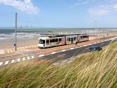 Strassenbahn De Lijn fährt auf Schienen entlang des Sandstrands von Raversijde bei blauem Himmel.