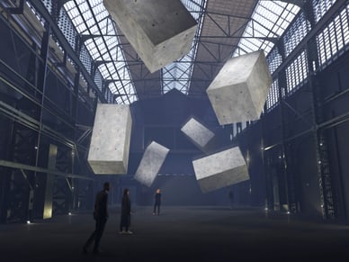 Drei Personen in einer Industriehalle mit schwebenden grauen Betonwürfeln unter einem Glasdach im Drift Museum Amsterdam.