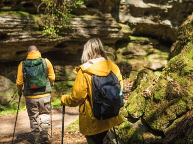 Mann und Frau mit Rucksäcken und Wanderstöcken wandern durch moosbedeckte Felsen im Wald.