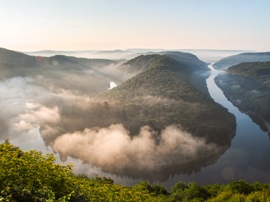 Luftaufnahme einer Flussbiegung mit bewaldeten Hügeln im Nebel.