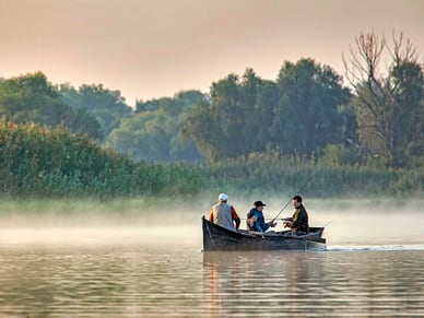 Drei Männer in einem traditionellen Fischerboot auf dem nebligen Donaudelta, umgeben von Schilf und Wald.