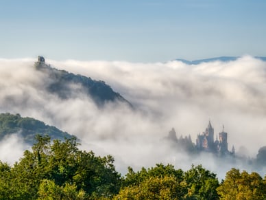 Zwei Schlossbauten im Morgennebel über dem Siebengebirge, umgeben von herbstlichem Waldgrün.