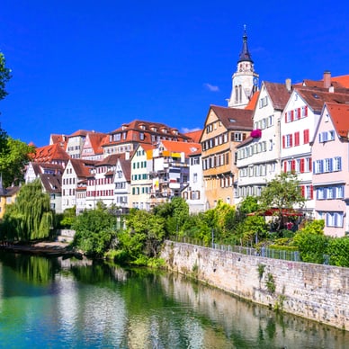 Bunte Altstadthäuser am Flussufer mit Kirchturm im Hintergrund.