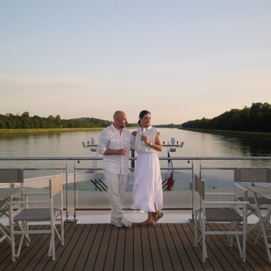 Zwei Personen in weisser Kleidung stehen auf dem Deck eines Bootes, umgeben von Tischen und Stühlen, mit Blick auf eine ruhige Wasserlandschaft.
