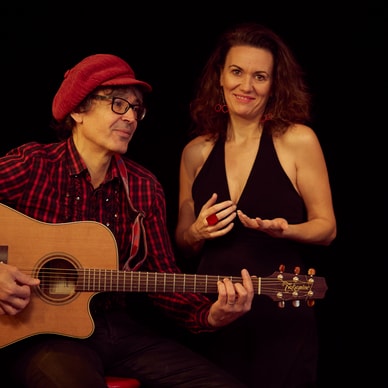 Person mit Gitarre und Frau in schwarzem Kleid auf schwarzem Hintergrund.