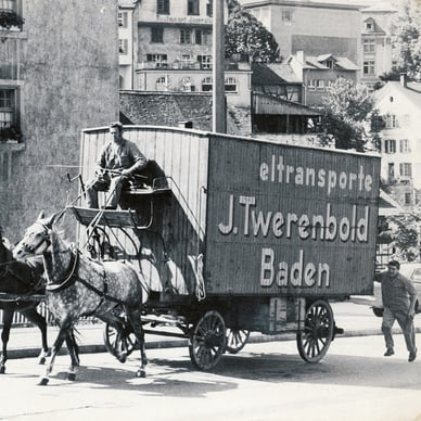 Schwarz-Weiss-Foto eines Pferdegespanns mit geschlossenem Wagen der Firma J. Twerenbold auf einer Strasse in Baden, Schweiz.