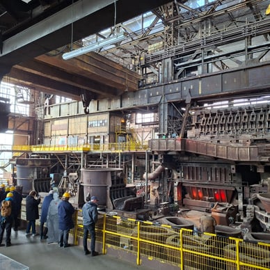 Menschen in gelben Helmen besichtigen eine industrielle Anlage mit Maschinen und Metallstrukturen.
