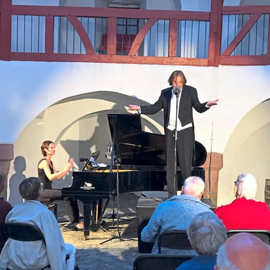 Musiker im Frack singt neben Pianistin auf Innenhofbühne vor Publikum.