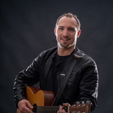 Mann in schwarzer Jacke mit Akustikgitarre vor dunklem Hintergrund.