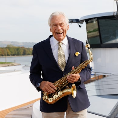 Pepe Lienhard im blauen Anzug hält ein goldenes Saxophon und lächelt auf dem Sonnendeck eines Flussschiffs.