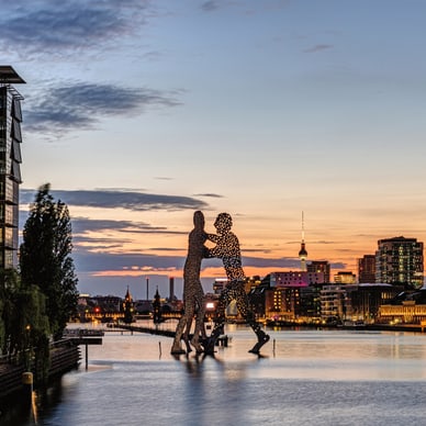 Zwei punktierte Silhouetten-Skulpturen spiegeln sich im Abendlicht in der Spree, dahinter der Berliner Fernsehturm.