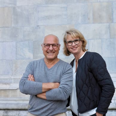 Eine lächelnde Frau und ein lächelnder Mann posieren vor einer grauen Steinmauer.