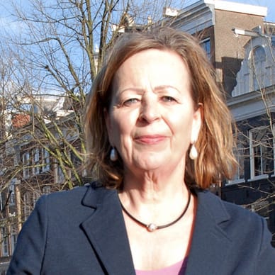 Frau mit dunklem Blazer steht vor einem Gebäude und Bäumen.
