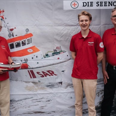 Drei Personen in roten Hemden stehen vor einem Bild eines Rettungsboots.
