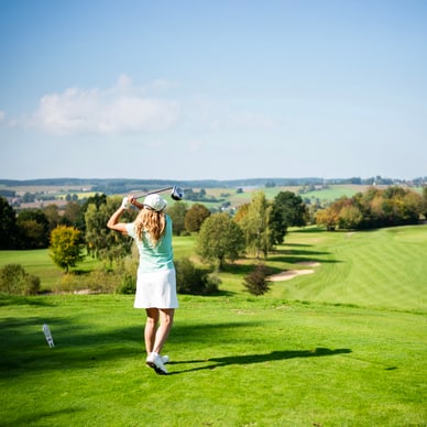 Frau in hellgrünem Shirt und weissem Rock führt einen Golfschlag auf einem weitläufigen Golfplatz aus.
