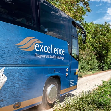Blauer Excellence-Bus am Flussufer, im Hintergrund das Flusskreuzfahrtschiff Excellence Baroness am Steg.