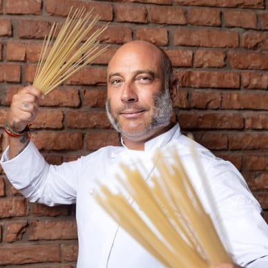 Mann in weisser Kochjacke hält Spaghetti vor einer Ziegelwand.