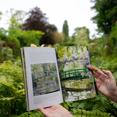 monets-seerosenteich-in-giverny.jpg Person hält ein offenes Buch mit Gartenbildern vor üppiger grüner Landschaft.