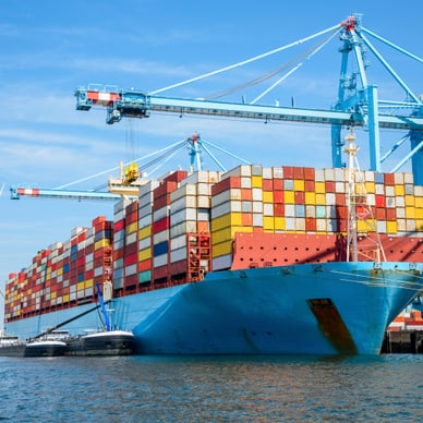 Grosses Containerschiff im Hafen mit blauen Kränen und Windkraftanlage im Hintergrund.