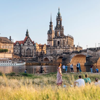 Personen entspannen im Gras mit Blick auf historische Gebäude und eine Brücke am Flussufer.