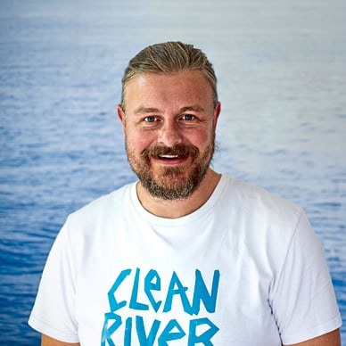 Mann im T-Shirt mit 'Clean River'-Aufdruck vor Wasserhintergrund.