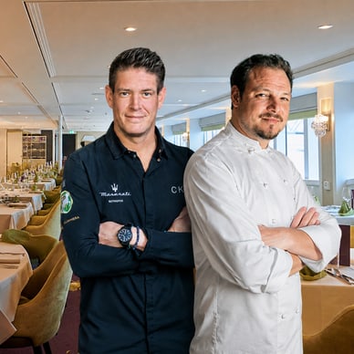 Zwei Männer in Kochjacken stehen in einem eleganten Restaurant mit gedeckten Tischen.