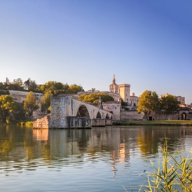 Steinerne Bogenbrücke Pont d'Avignon spiegelt sich im Rhône, dahinter der historische Papstpalast unter blauem Himmel.