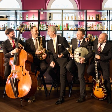 Fünf Männer in Anzügen mit Musikinstrumenten vor einer Bar.
