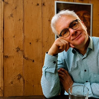 Älterer Mann mit Brille sitzt an einem Holztisch.