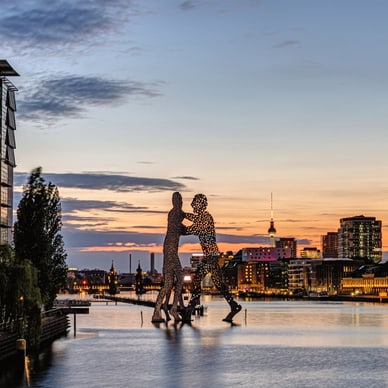 Zwei punktierte Silhouetten-Skulpturen spiegeln sich im Abendlicht in der Spree, dahinter der Berliner Fernsehturm.